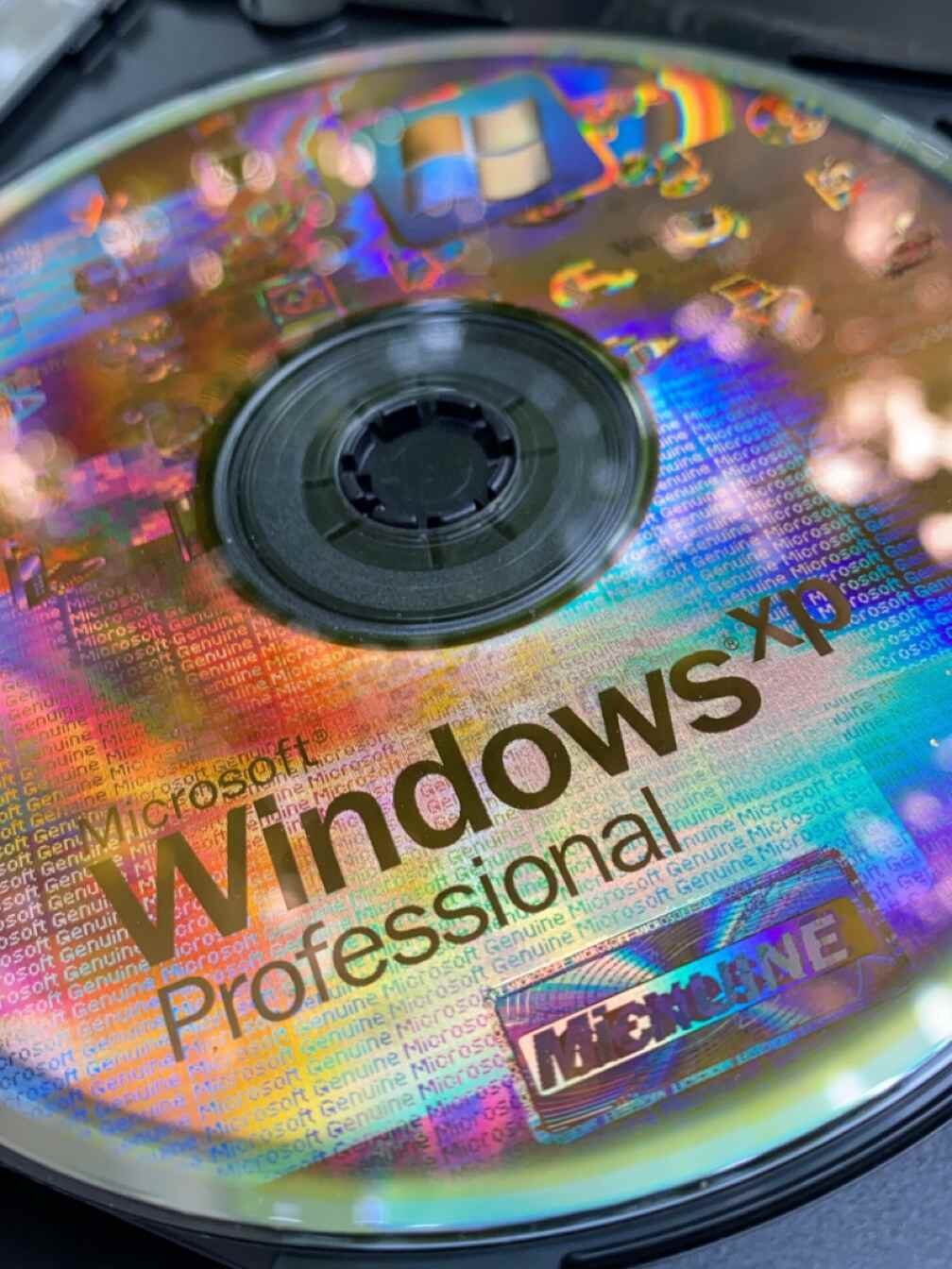 A silver Windows XP Professional installation CD sitting in an open optical drive, its surface showing rainbow holographic anti-counterfeiting patterns and the words Microsoft Windows XP Professional.