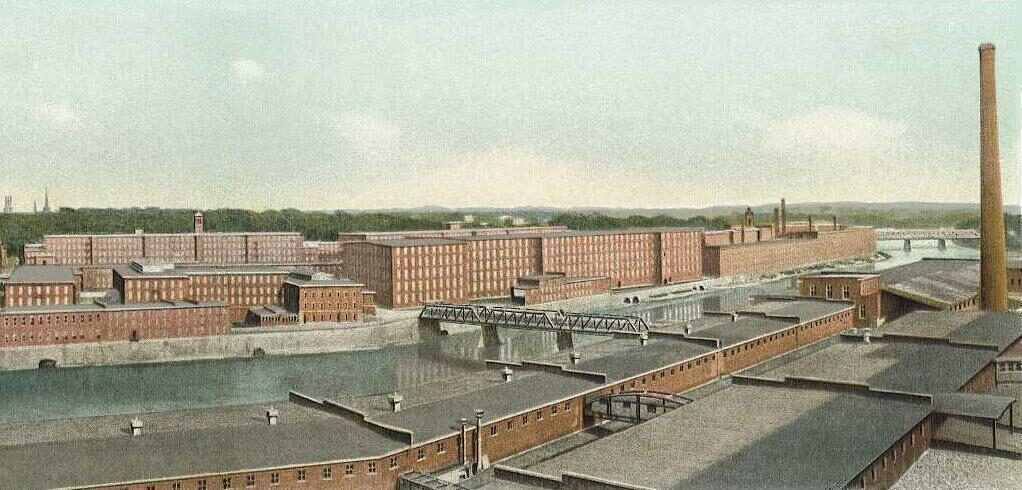 A colour-tinted postcard panorama of long multi-storey brick textile mills lining a river and its canals, with a truss bridge and a dam in the middle distance and a tall chimney at the right.