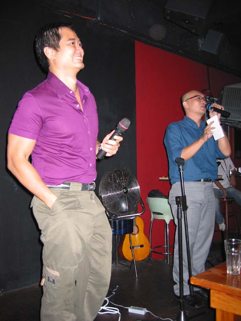 Two men with microphones stand in a small dark venue with a black and red wall; one in a purple shirt smiles broadly, the other, in a blue shirt and glasses, speaks from a card.