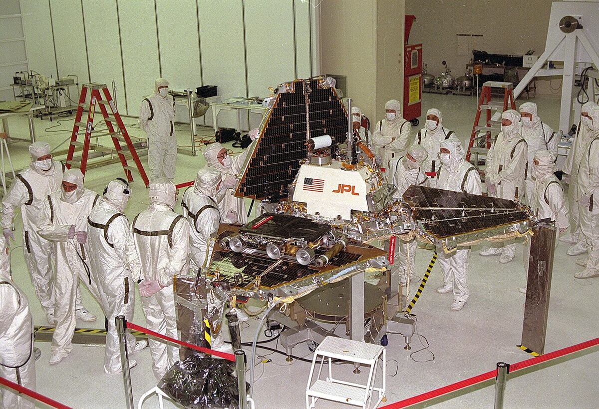 A cleanroom full of people in white coveralls and masks standing around a three-petalled spacecraft lander with solar panels, a small six-wheeled rover on one petal.