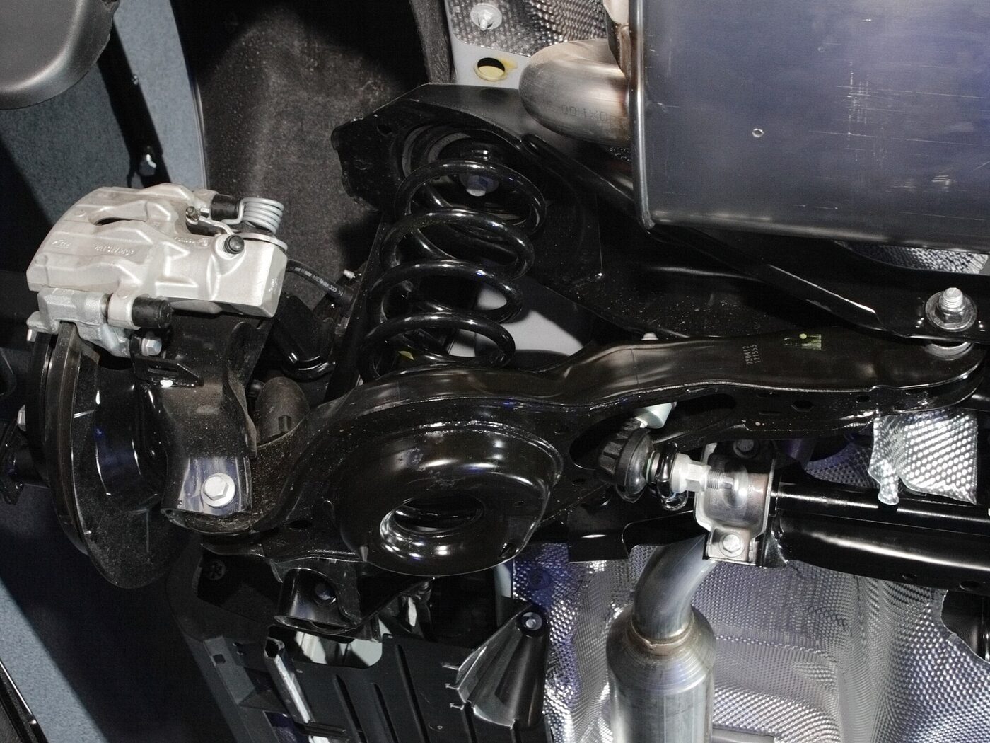 Close-up from below of a car's rear suspension corner, showing a broad flat pressed-steel arm running to the wheel hub and brake caliper, a black coil spring, tubular lateral links and an exhaust silencer alongside a heat-shielded floorpan.