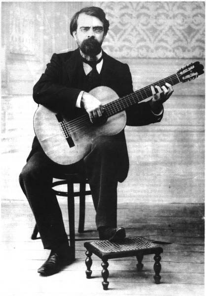 Black and white studio photograph of a seated bearded man in a dark suit playing a classical guitar, with a small wooden footstool on the floor in front of him