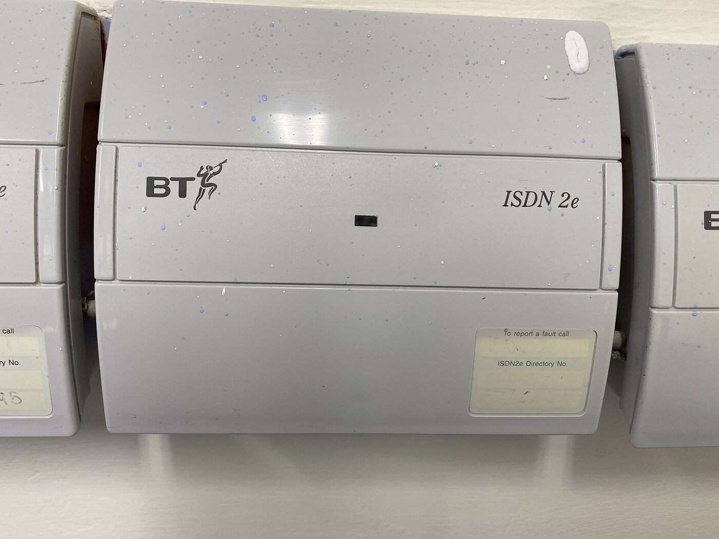 Close-up of a grey plastic wall box marked BT and ISDN 2e, with an unlit indicator window and a fault-reporting label, flanked by two identical boxes.