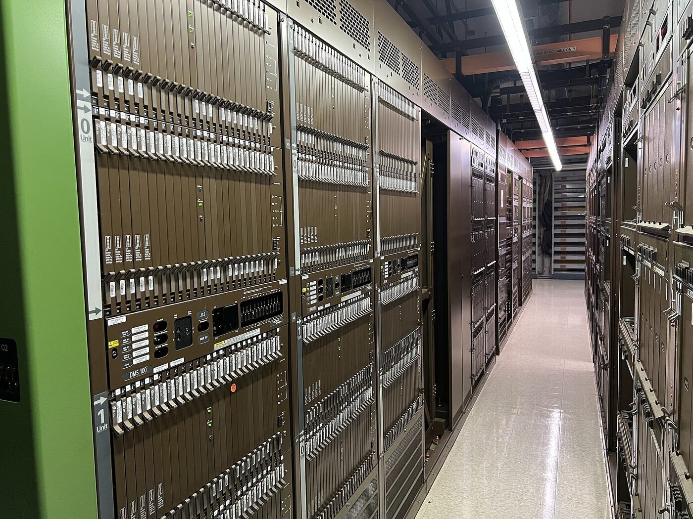 A narrow aisle between tall racks of telephone switching equipment in a central office, the frame nearest the camera labelled DMS 100 and capped in green.