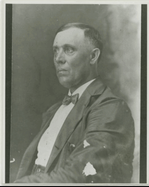 Black-and-white studio portrait of a seated middle-aged man in a dark suit and bow tie, turned slightly away from the camera.