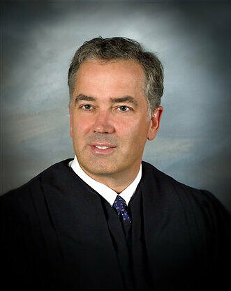 Formal portrait photograph of a man in black judicial robes with a dark patterned tie, against a grey studio background.