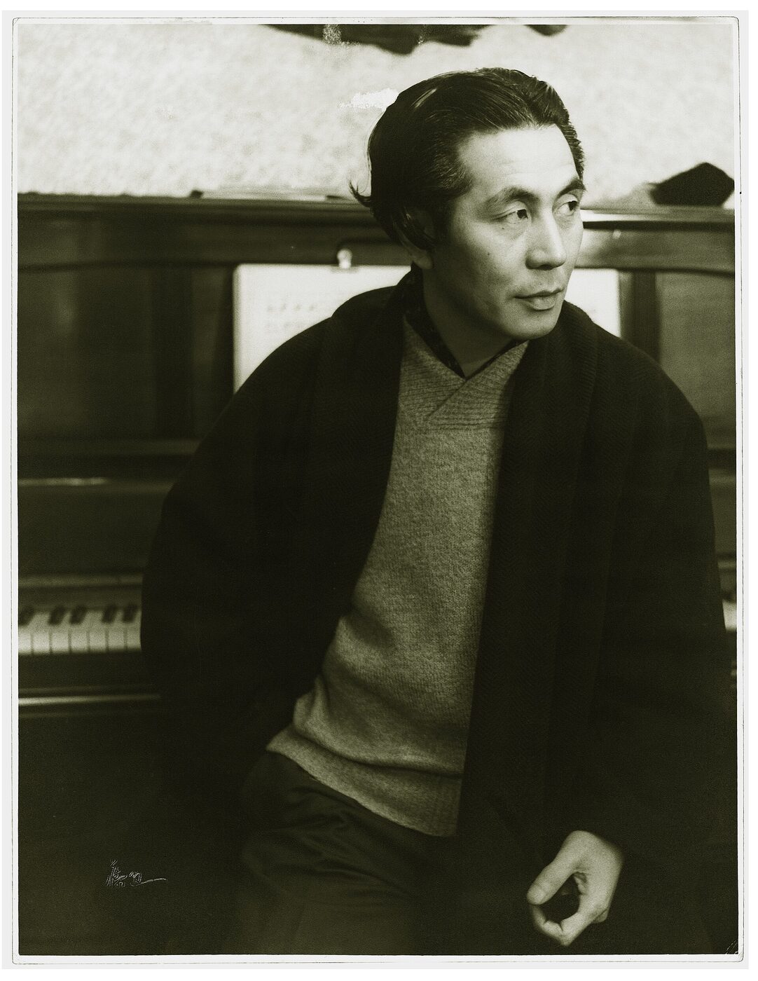 Black-and-white portrait of a seated man in a sweater and dark jacket leaning against an upright piano.