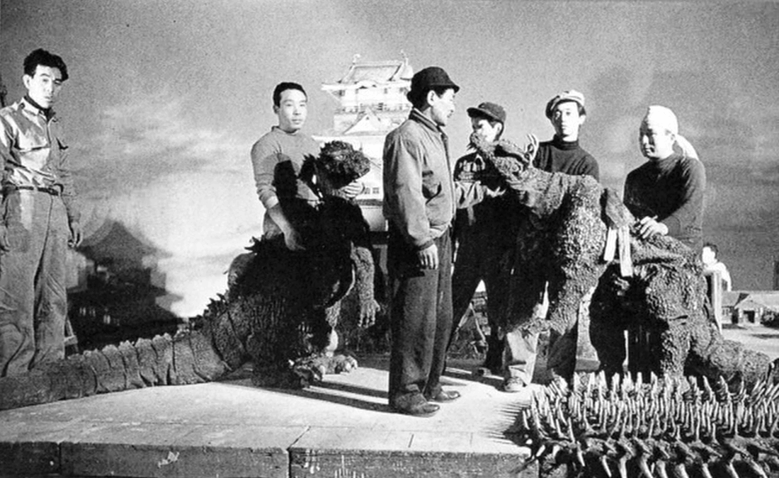 Black-and-white photograph of two monster costumes supported by film crew on a studio stage, with a model castle behind them.