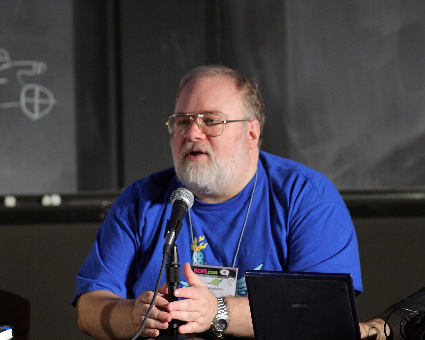 A bearded man in a blue T-shirt and a conference badge speaking into a microphone at a panel table, a laptop beside him.