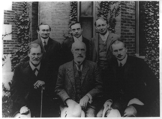 A formal black-and-white group photograph of six men in dark suits, three seated in front and three standing behind, posed outdoors against a plain background.