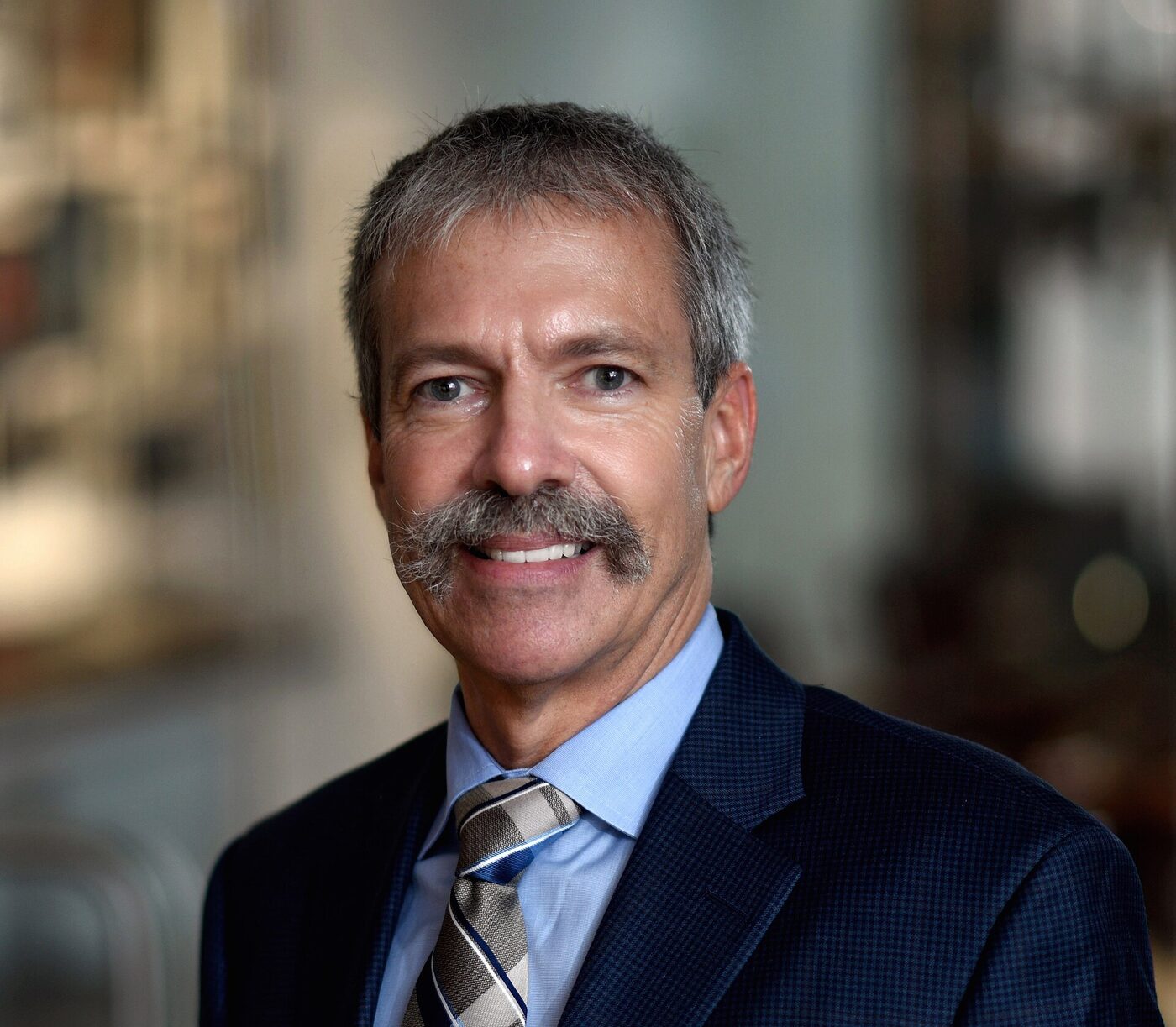 Studio portrait of a smiling grey-haired man with a moustache, wearing a dark jacket, pale blue shirt and patterned tie.