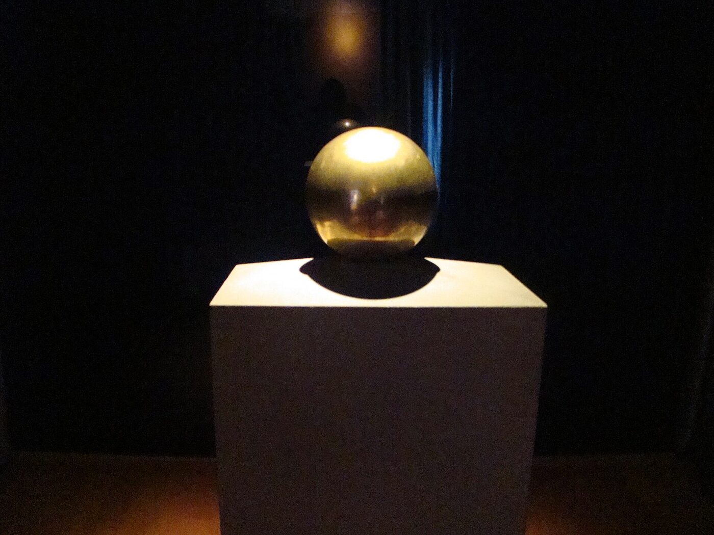 A polished gold-coloured sphere resting on a pale rectangular pedestal in a darkened museum room, lit from above, with the surrounding wall in shadow.