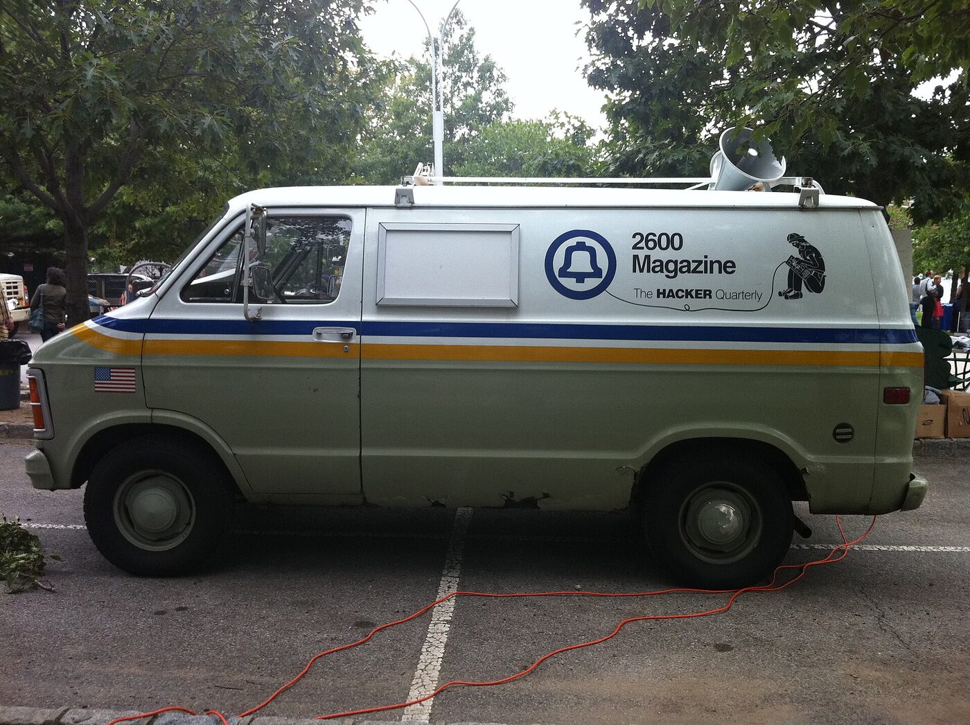 A white utility van in telephone-company livery, its lettering altered to read as a hacker magazine's name, parked on grass at an outdoor event.