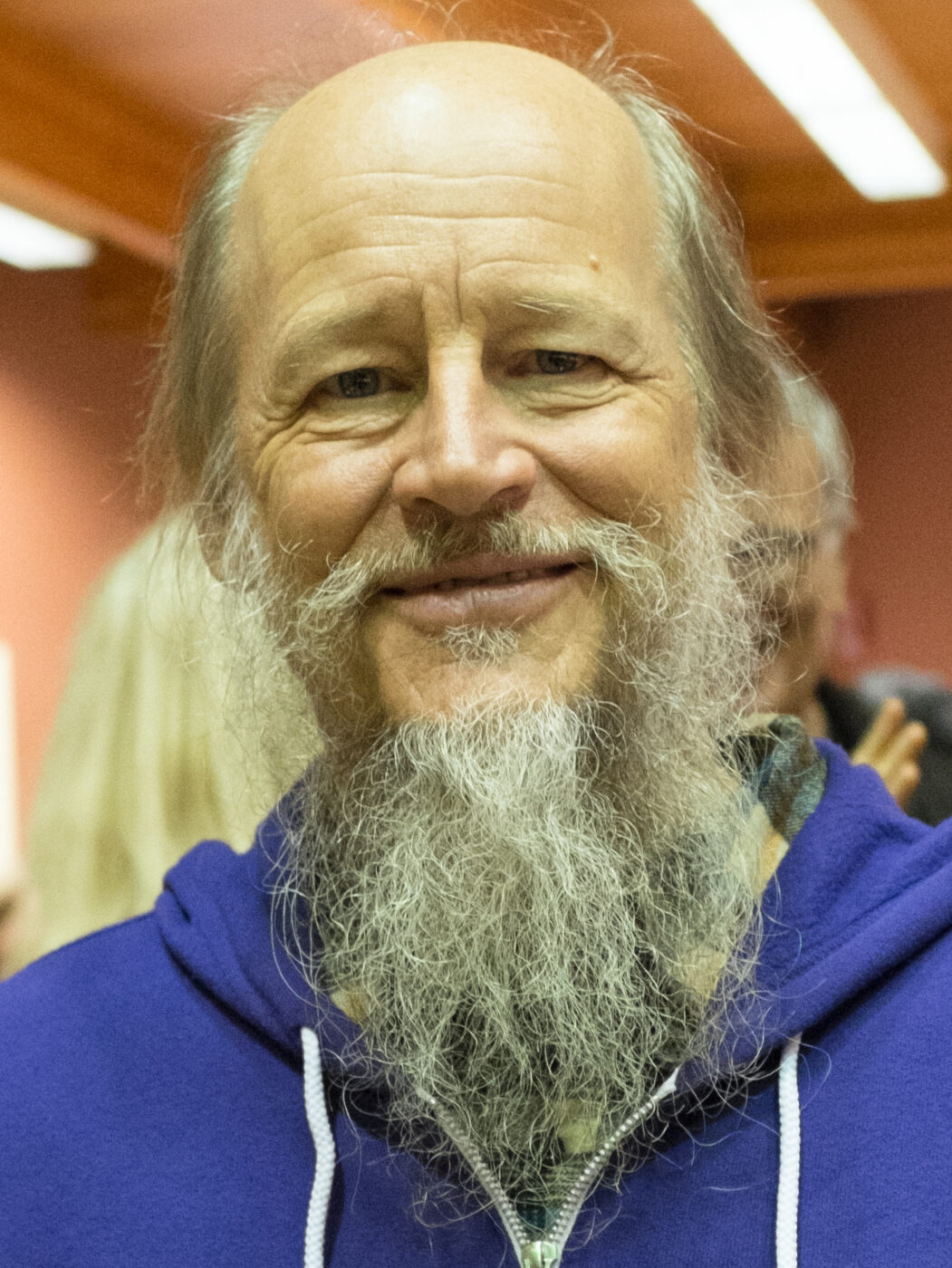 Head-and-shoulders photograph of a smiling older man, bald on top with long grey hair and a long grey beard, wearing a blue hooded sweatshirt indoors.