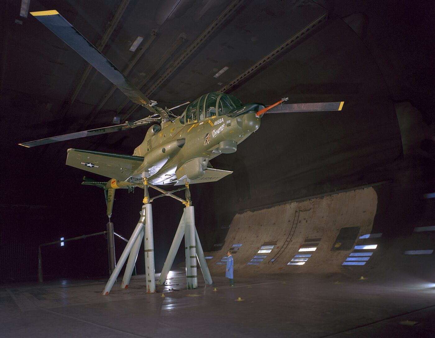 A large attack helicopter with a rigid rotor and stub wings, mounted on supports inside a cavernous wind tunnel test section.