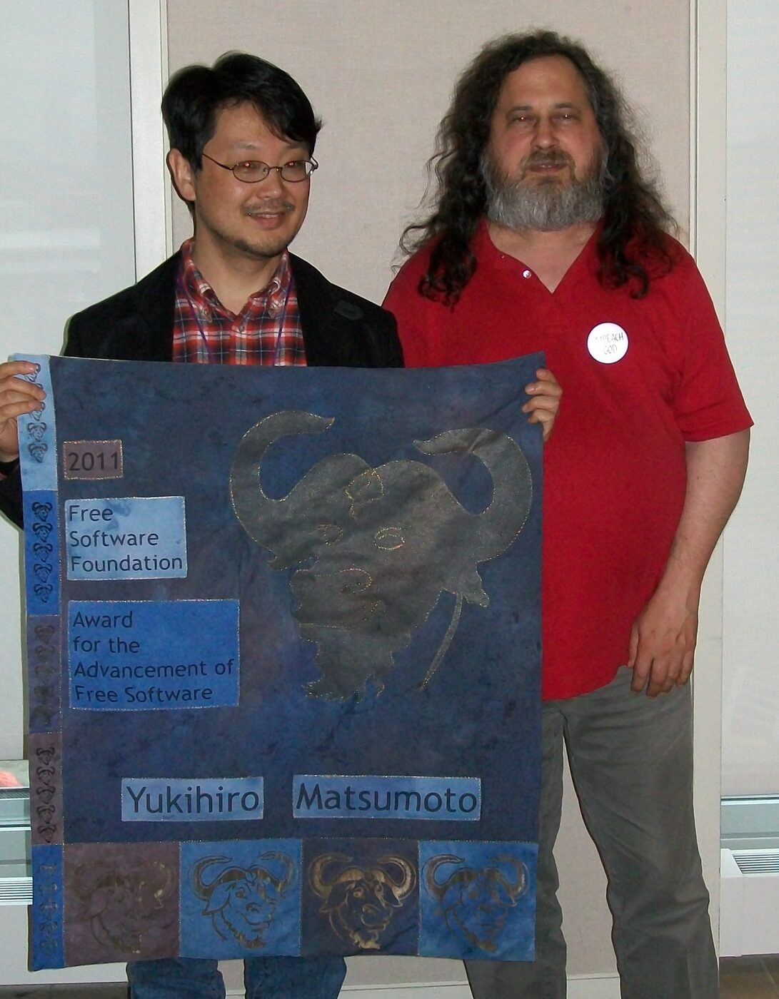 Two men standing side by side holding a large quilted banner bearing a gnu head and the words Free Software Foundation Award for the Advancement of Free Software
