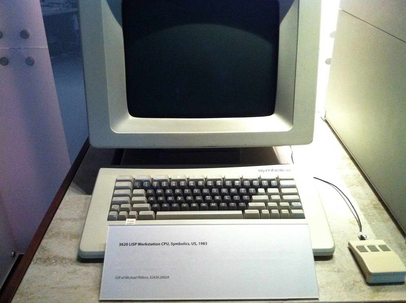 A beige 1980s Symbolics workstation with a large monitor, keyboard and mouse, displayed with a museum label in an exhibit case.