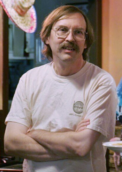 A man with shoulder-length hair, large metal-framed glasses and a moustache, arms folded, wearing a white T-shirt in a room among other people.