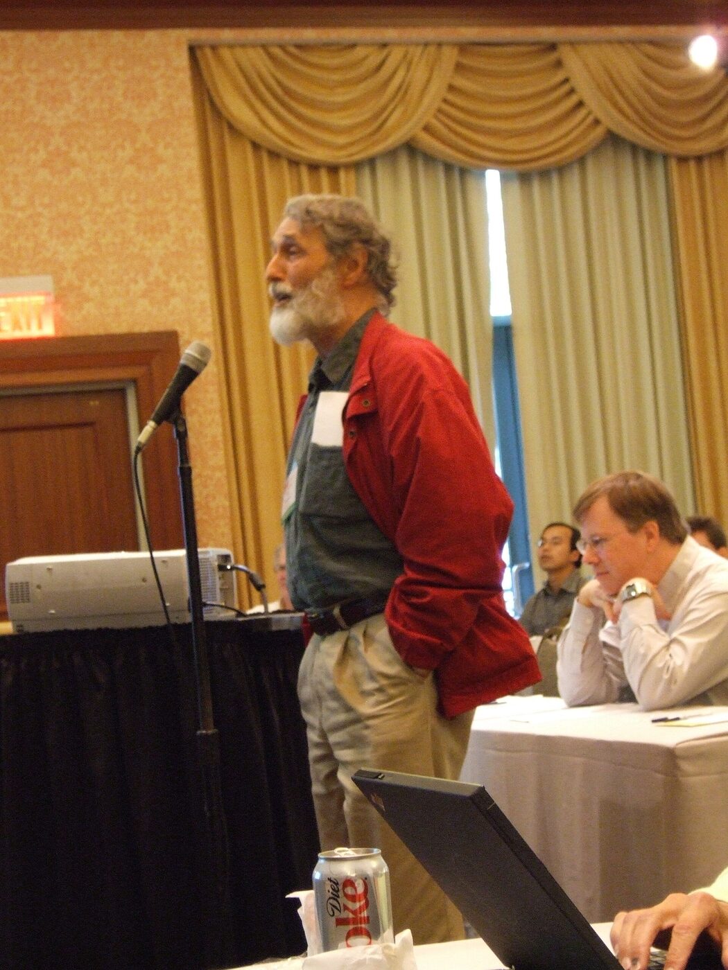 A white-bearded man in a red shirt and pale trousers standing at a floor microphone in a hotel conference room, with seated attendees at draped tables behind him.