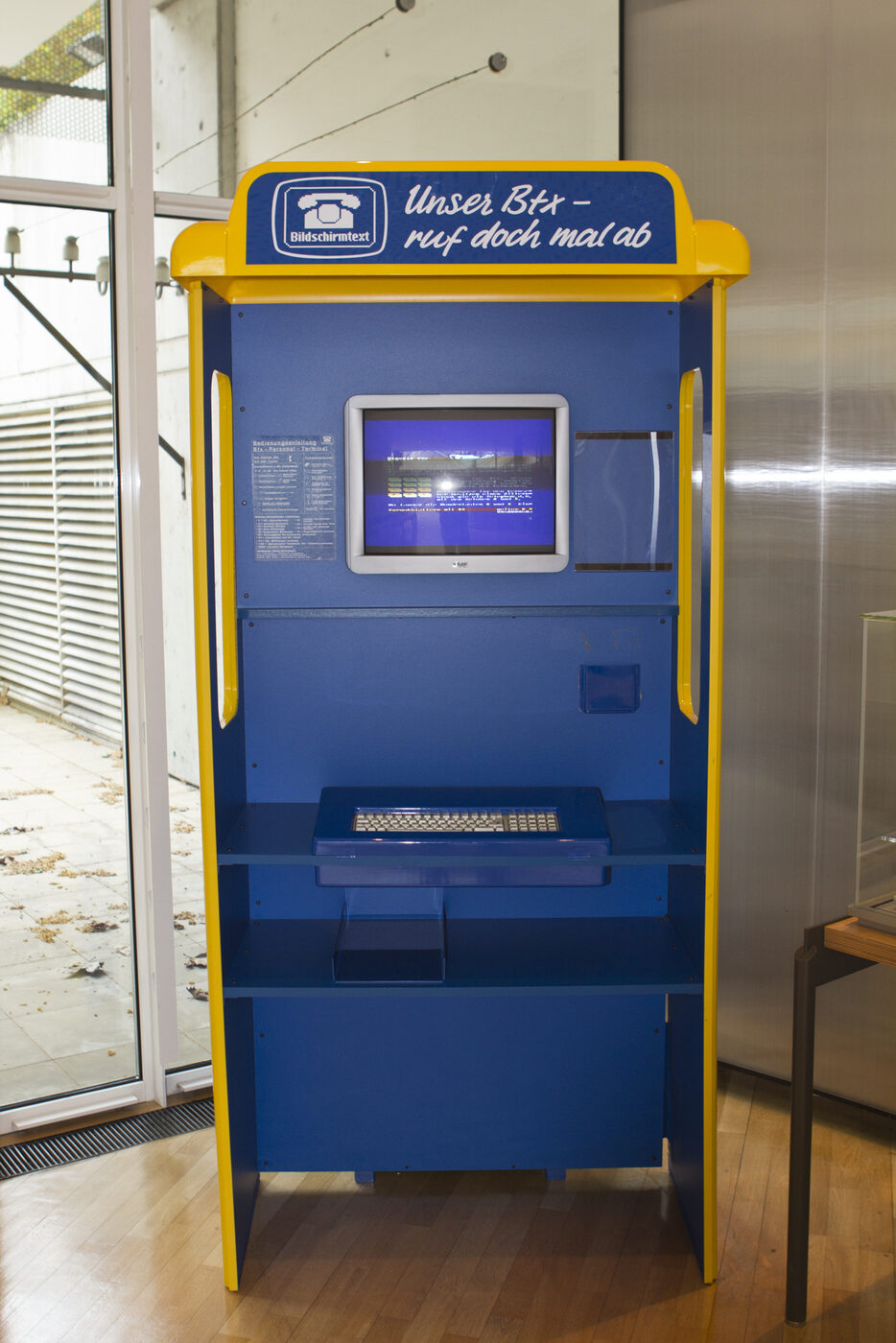 A free-standing public Bildschirmtext terminal in blue and yellow Bundespost livery: a small CRT screen set into the upper panel above a full typewriter keyboard on a pull-out shelf, photographed in a museum display.