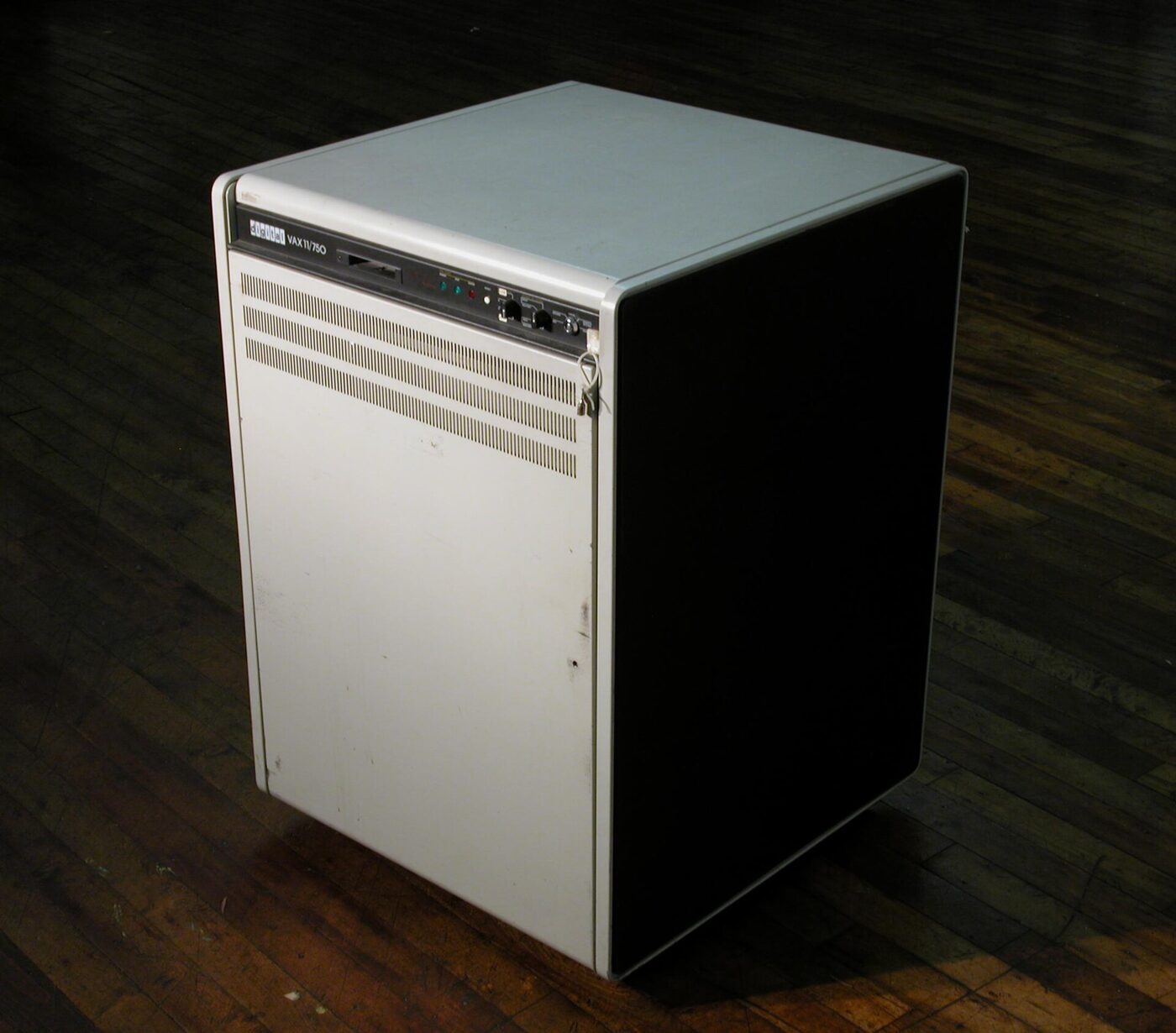 A squat off-white and black minicomputer cabinet lettered "digital VAX 11/750", with a small control panel and slot on the front, standing on bare floorboards.