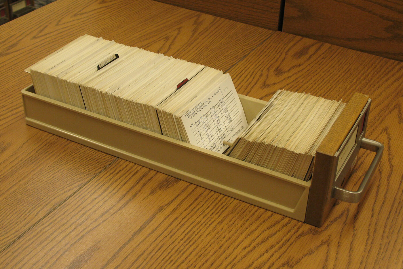 A card-index drawer lifted out of its cabinet and set on a library table, packed with typed catalogue cards, one card raised upright to be read.
