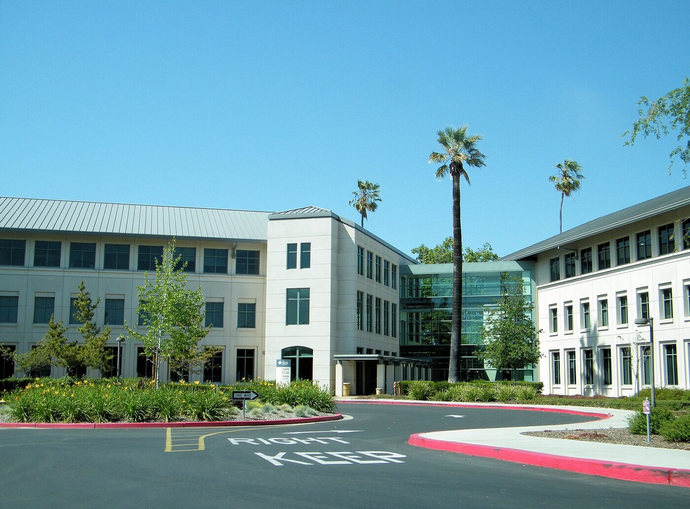 Two three-storey cream-coloured office buildings joined by a glass atrium, with palm trees and a landscaped roadway in front.