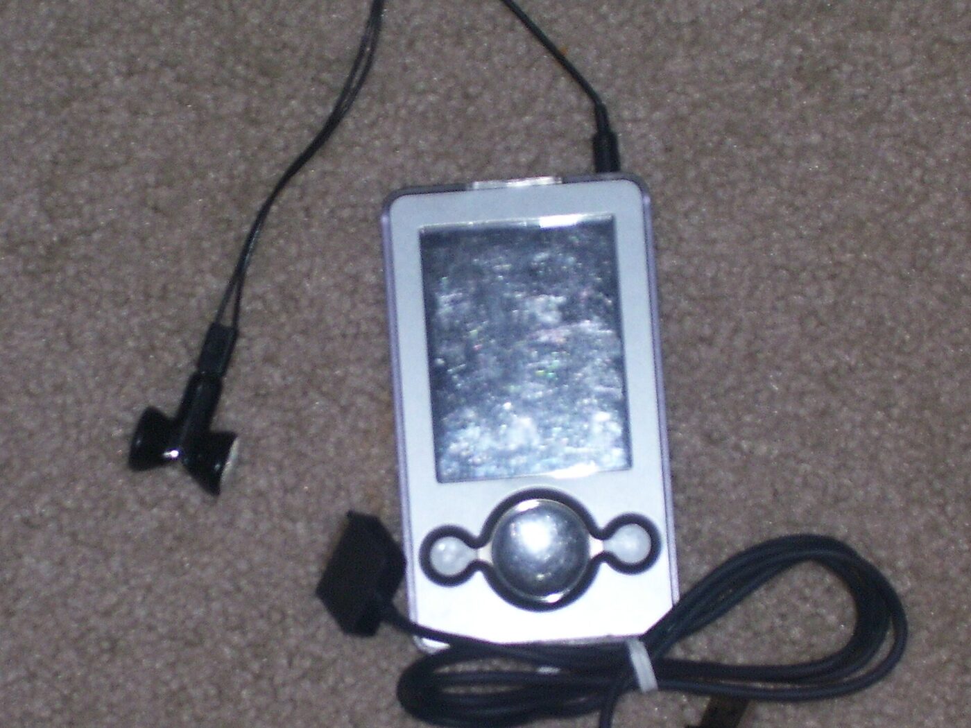 A silver-and-white portable media player lying face up on beige carpet, with a rectangular screen above a round control pad flanked by two small buttons, a black earphone lead running away to one side and a coiled black sync cable beside it.