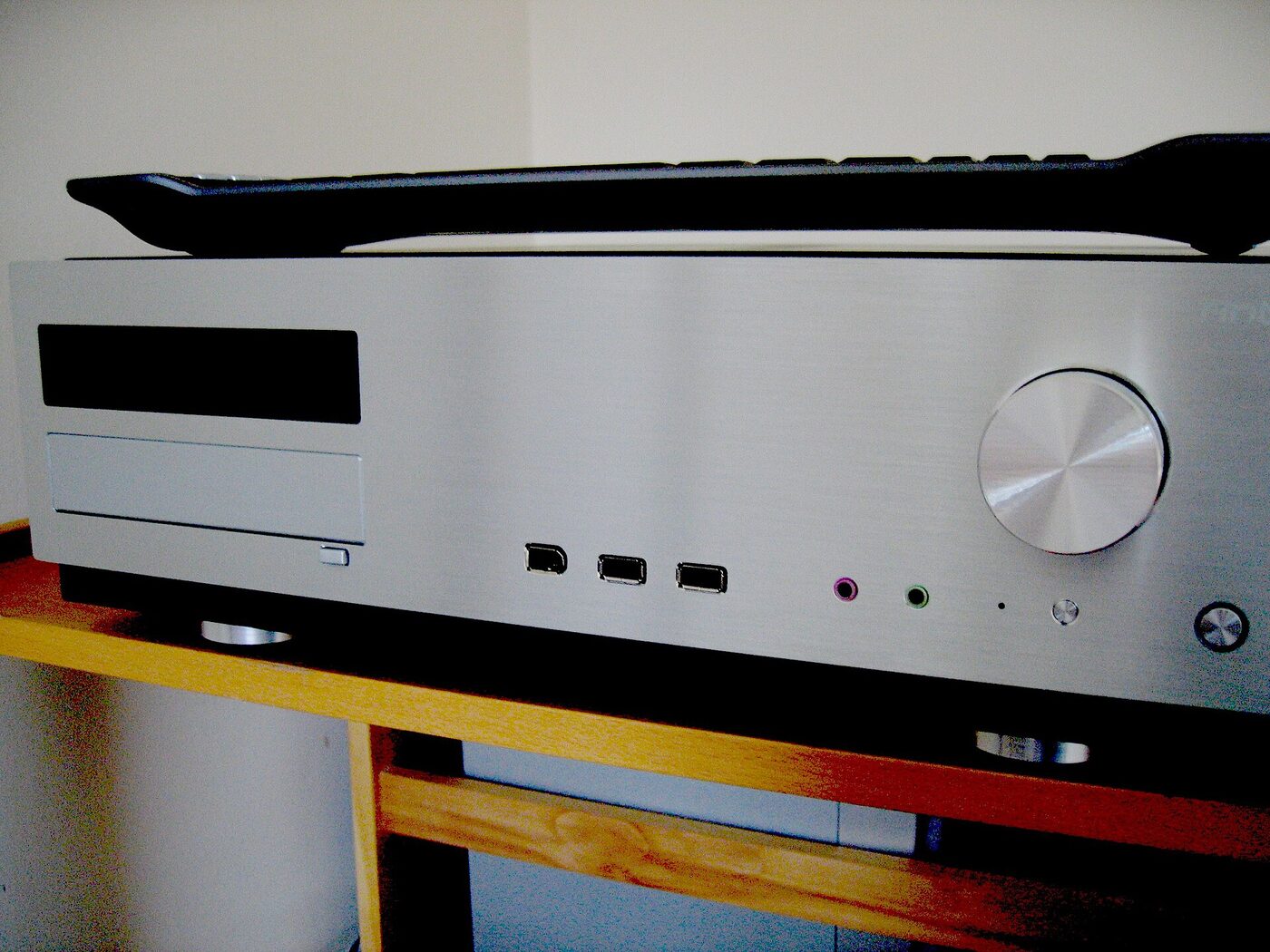 A slim silver hi-fi-style computer case standing on a wooden rack, with a volume knob, three front USB sockets, headphone and microphone jacks and a dark display panel; a black keyboard lies on top of it.
