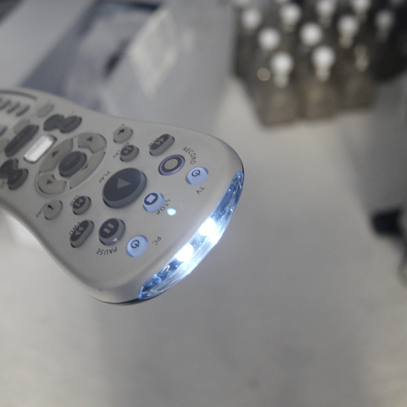 Close-up of a pale grey handheld remote control lying on a desk, its emitters at the narrow end glowing bright white, with buttons marked play, pause, stop and record visible on the keypad.