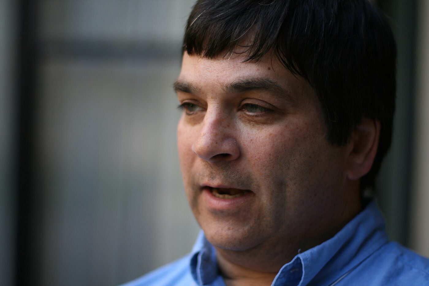 Close-up photograph of a dark-haired man in a blue shirt, caught mid-sentence.