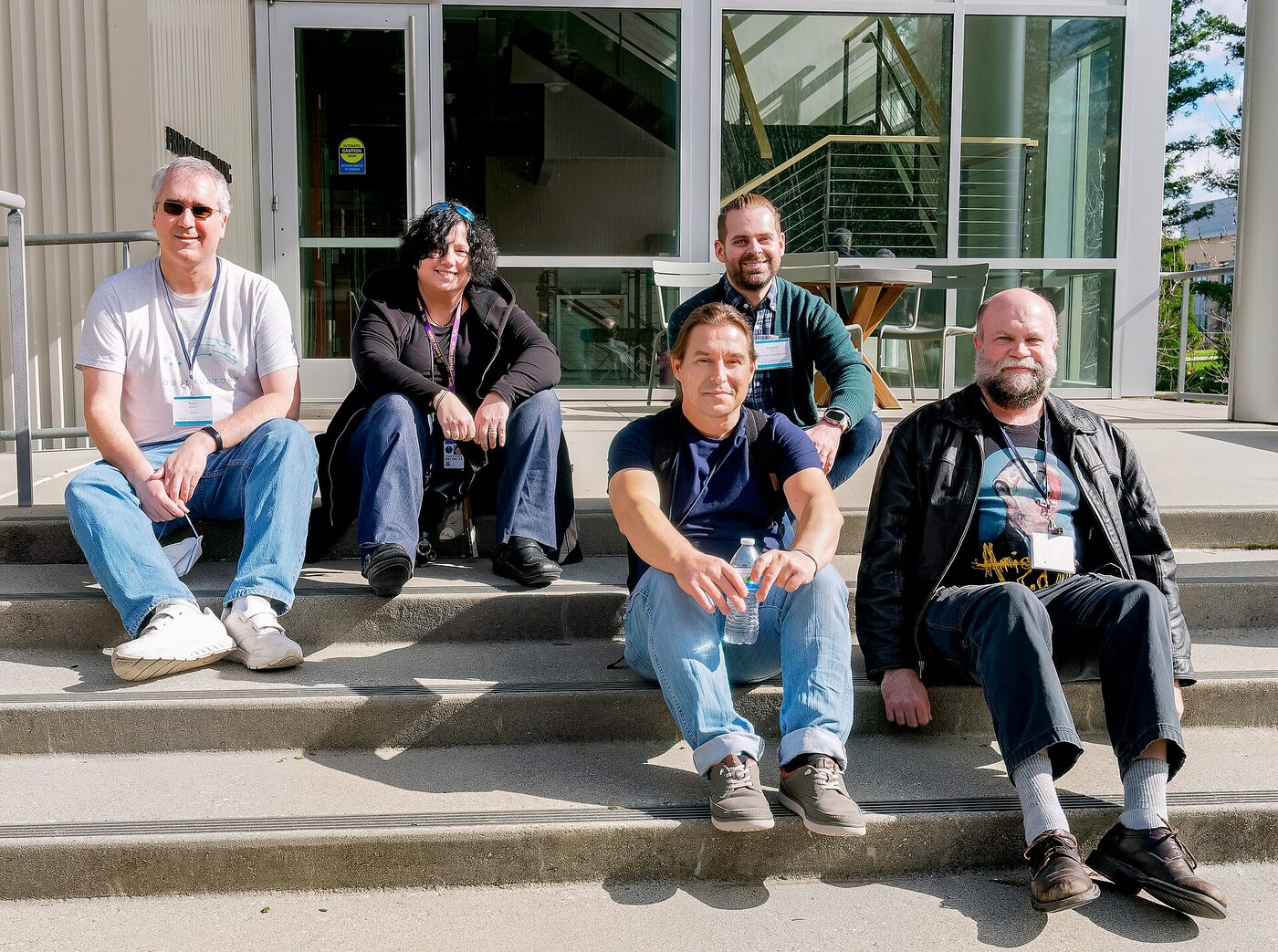 Five people sitting on concrete steps outside a modern glass-fronted building, facing the camera.