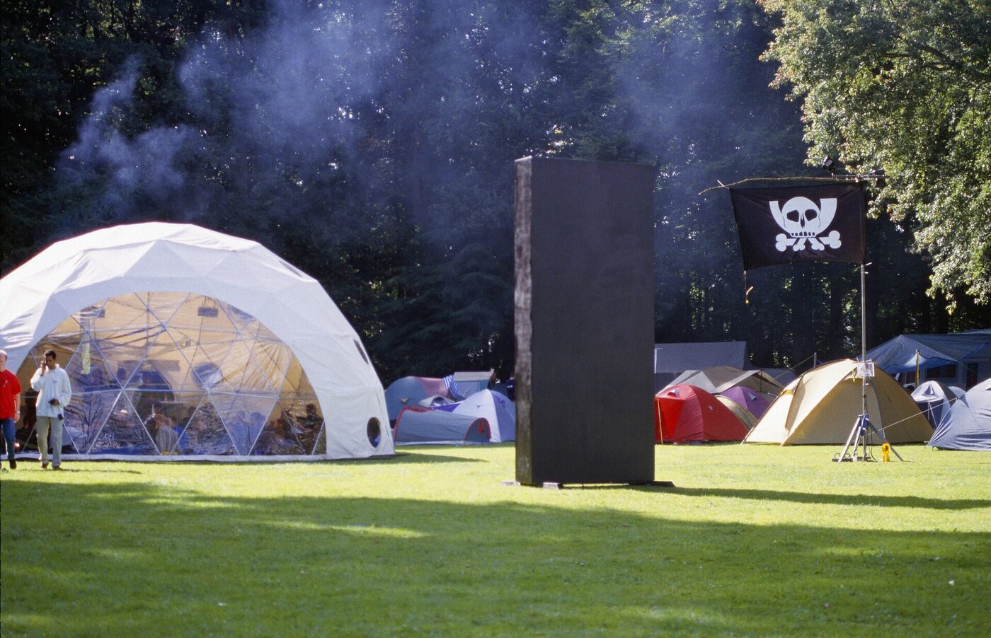 A tall black rectangular slab standing on a grass field among dome and ridge tents, with a skull-and-crossbones flag flying beside it and woodland behind.