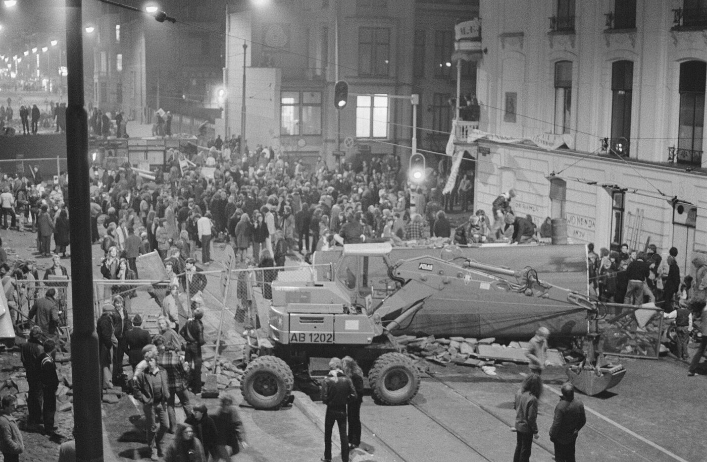 Black-and-white night photograph of a crowded Amsterdam street with tram rails, torn-up paving stones piled in the road and a mechanical excavator standing among the crowd.