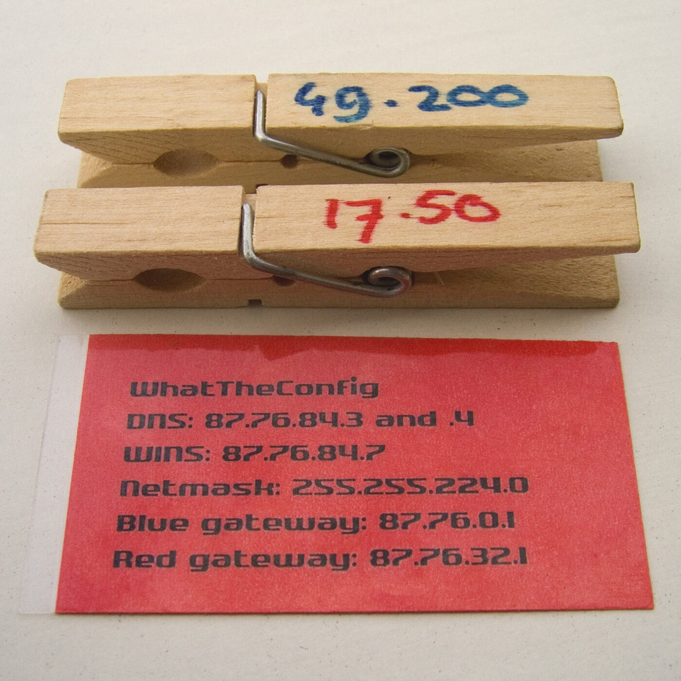 Two wooden clothes pegs, each hand-written with a pair of numbers, lying above a red printed card headed WhatTheConfig listing DNS, WINS, netmask and gateway addresses.