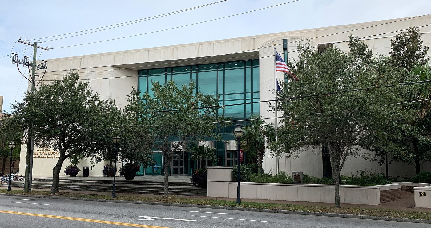 A long modern pale-stone library building with a tall green-tinted glass entrance bay, a flagpole flying the United States flag, and street trees along the pavement in front, photographed from across the road.