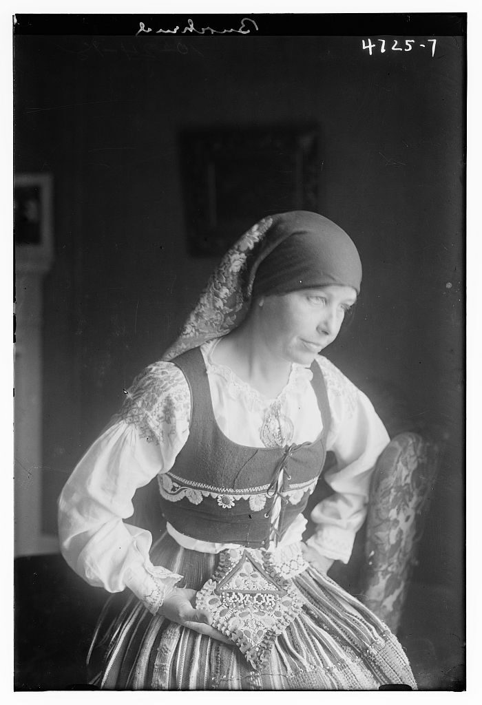 Black and white studio photograph of a woman seated indoors, turned to one side, wearing an embroidered bodice over a full white blouse, a patterned apron and a headscarf edged with lace.