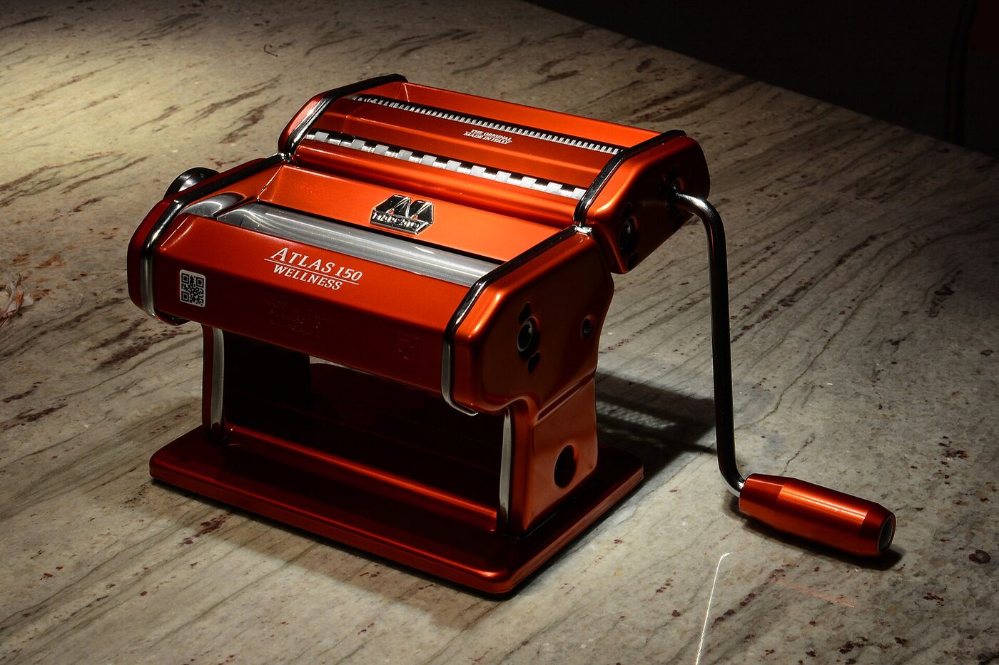 A red enamelled hand-cranked pasta machine marked ATLAS 150 WELLNESS, with smooth rollers below and a toothed cutting roller above, its crank handle fitted into the side, standing on a pale marble surface.