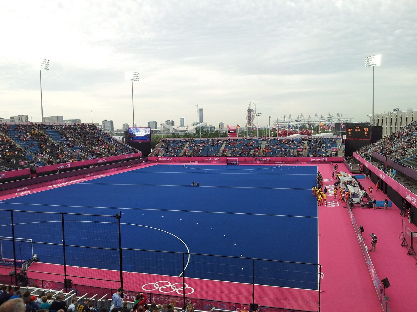 A blue synthetic hockey pitch with a bright pink surround seen from raised seating, with floodlights, temporary stands, a scoreboard reading ESP 0 AUS 0 and the Olympic Park skyline behind.