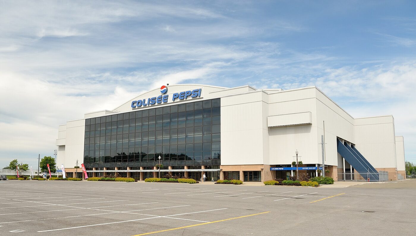 Exterior of a large pale arena with an arched roofline and a tall glass frontage, lettered Colisée Pepsi, seen across an empty car park under a blue sky.