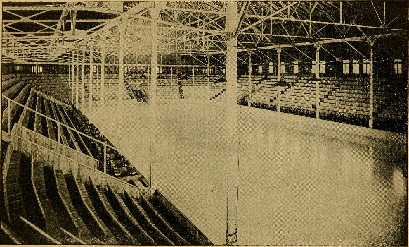Sepia halftone photograph of an empty indoor ice rink, with tiered bench seating rising on both sides and a lattice of steel roof trusses and slender columns overhead.