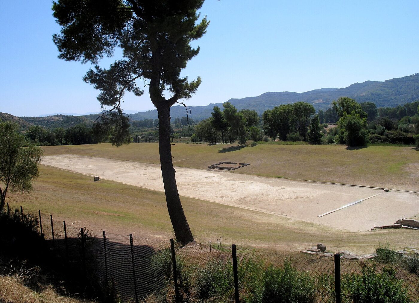 The bare earth track of the ancient stadium at Olympia, seen across the grassed embankments where spectators sat