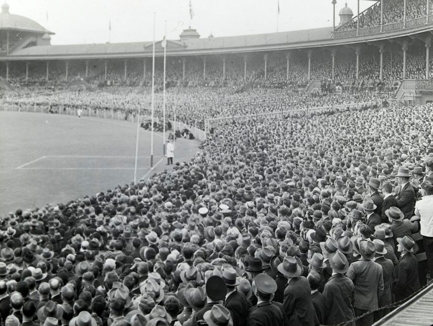 Black and white photograph of a dense hatted crowd filling the terraces and stands of the Melbourne Cricket Ground, with goal posts and part of the playing field at the left.