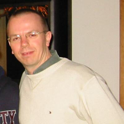 A man in glasses and a pale jumper turning towards the camera in a dimly lit room at a party.