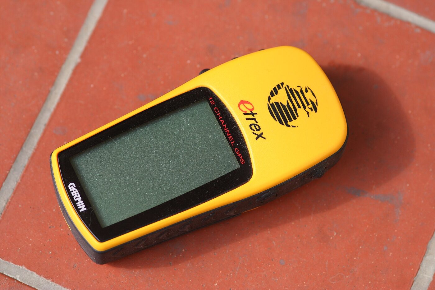 A yellow handheld GPS receiver lying face up on red floor tiles, its monochrome screen blank, with Garmin, eTrex and &ldquo;12 channel GPS&rdquo; markings visible on the case.