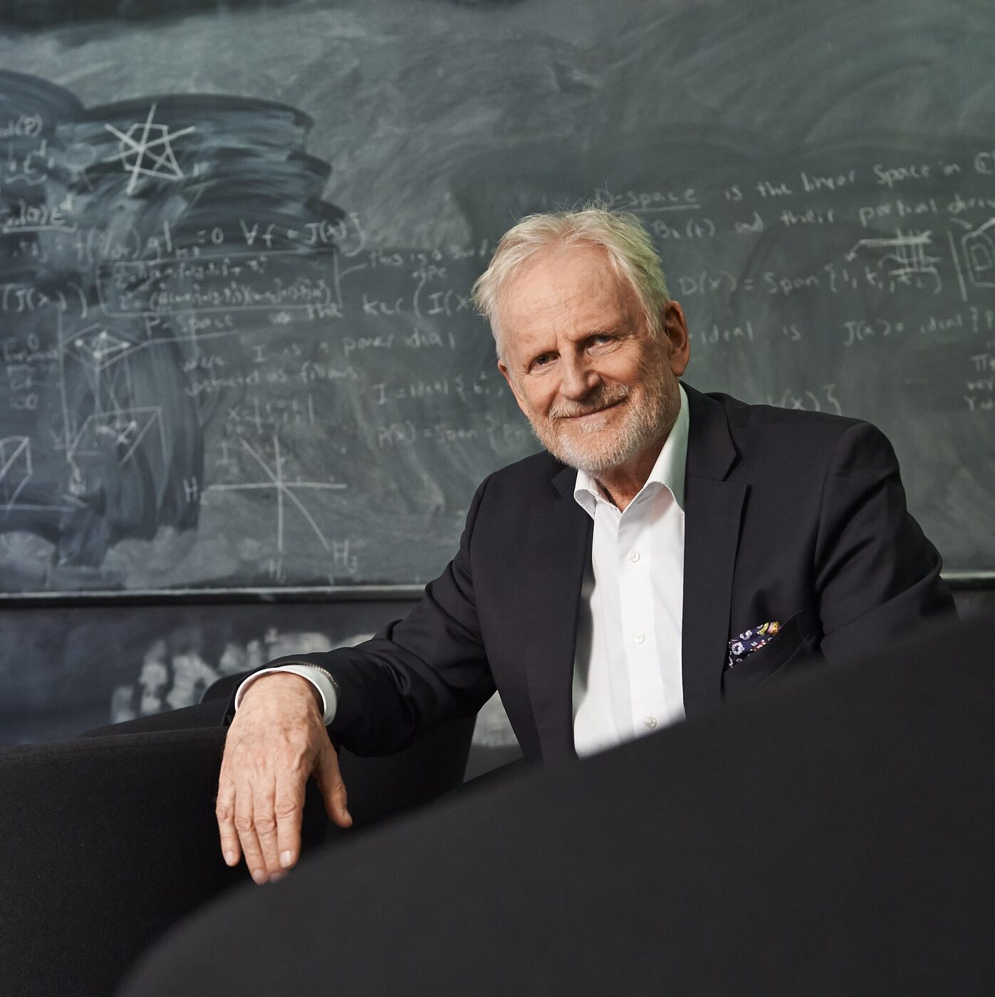 An elderly white-haired man in a dark jacket, leaning on the back of a chair, in front of a blackboard covered with handwritten algebra.