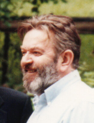 Colour photograph of a smiling, grey-bearded man in a light open-necked shirt, taken outdoors in front of greenery.
