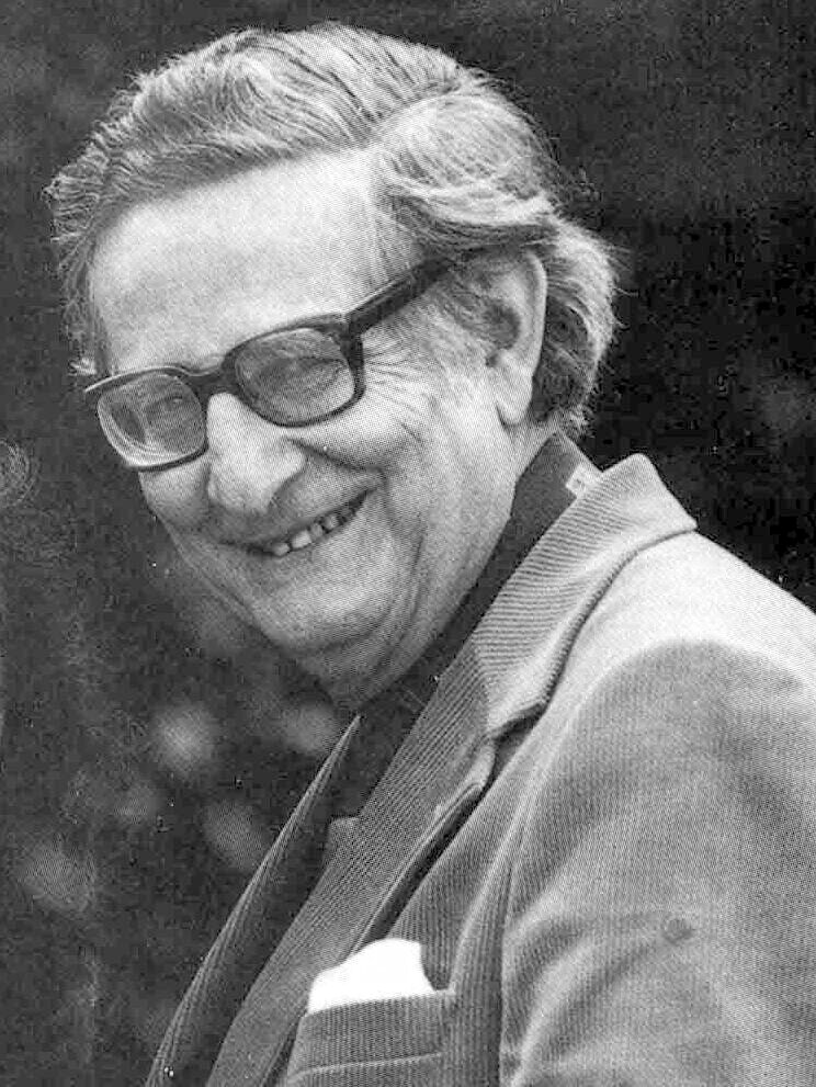 Black and white photograph of an older man with swept-back grey hair, wearing dark-rimmed glasses and a jacket, smiling and turned slightly away from the camera.