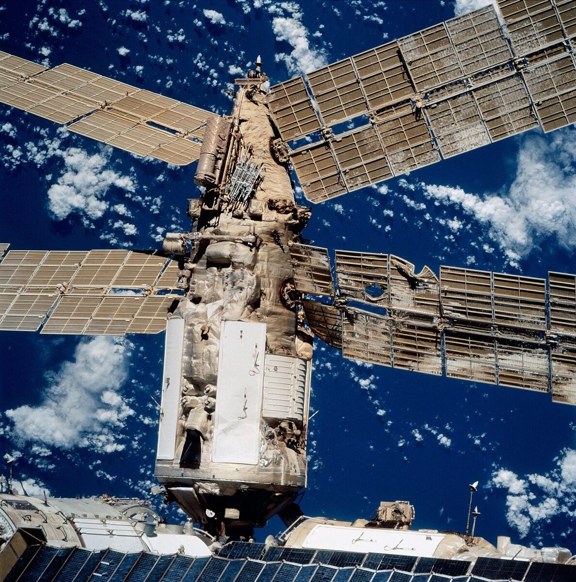 Close view of a space station module with a buckled and torn solar array and crumpled thermal insulation on the module’s exterior.
