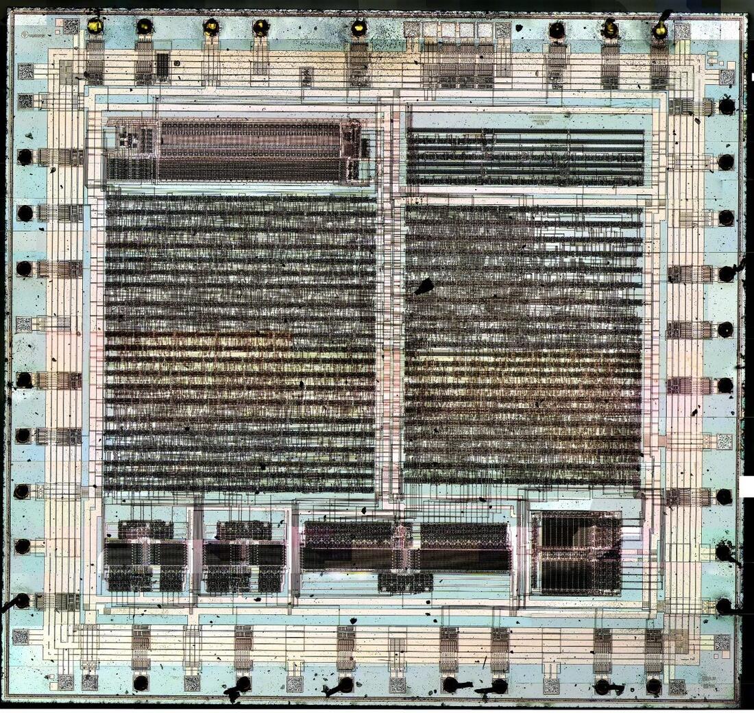 Colour micrograph of an exposed integrated-circuit die: a square of pale blue and tan silicon covered in dense rows of circuitry, with bond pads and wire stubs around all four edges.